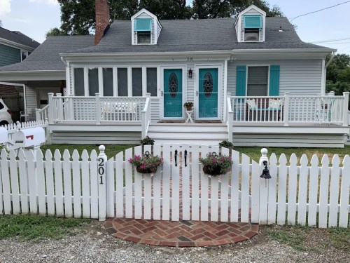 A charming white house with teal accents, a front porch, and a picket fence, featuring flower beds and a brick pathway.
