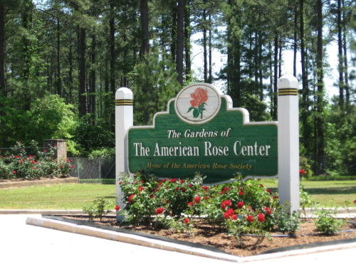 Sign for The Gardens of The American Rose Center, surrounded by greenery and blooming rose bushes.