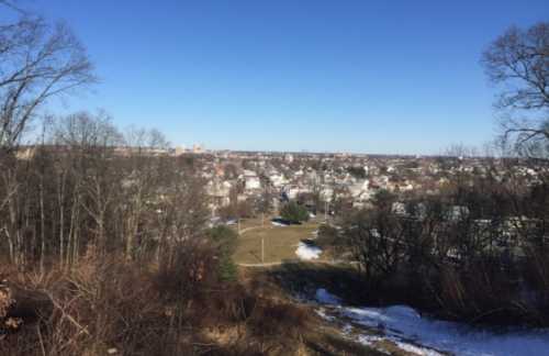 A panoramic view of a town from a hill, featuring houses, trees, and a clear blue sky. Snow is visible on the ground.