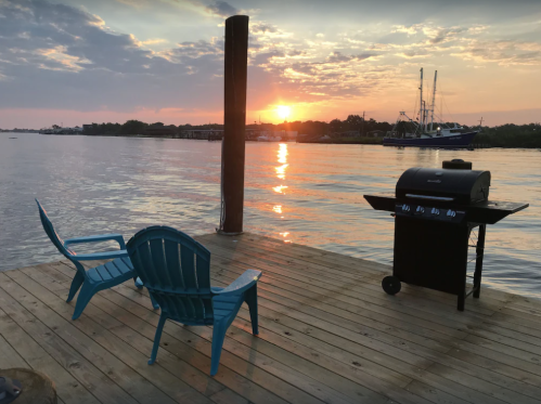 A serene sunset over water, featuring a grill and two blue chairs on a wooden dock.