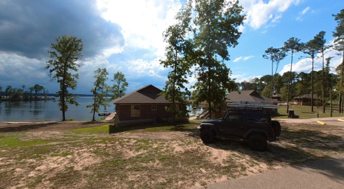 A lakeside scene featuring a house, a parked black Jeep, and trees under a partly cloudy sky.