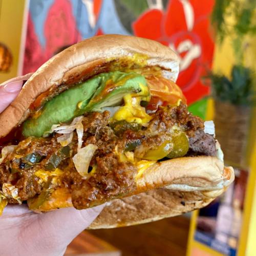 A close-up of a hand holding a loaded burger with avocado, cheese, and toppings against a colorful background.