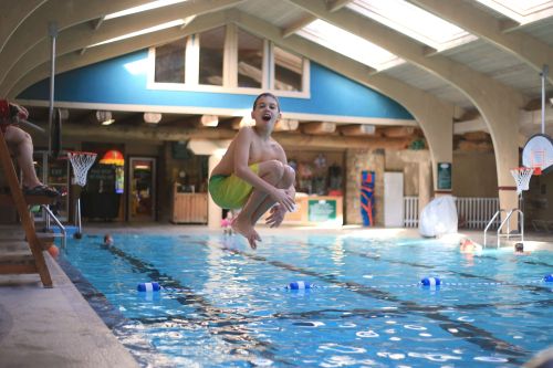 A child joyfully jumps into a swimming pool, surrounded by other swimmers and a bright indoor setting.