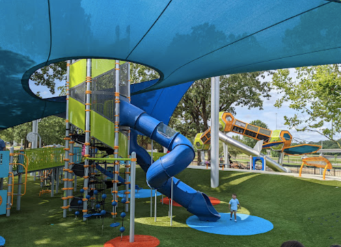 A colorful playground with a blue slide, climbing structures, and shaded areas under a blue canopy. Children playing nearby.
