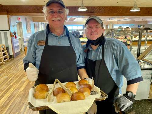 Two smiling bakers hold a tray of freshly baked rolls in a cozy dining setting.