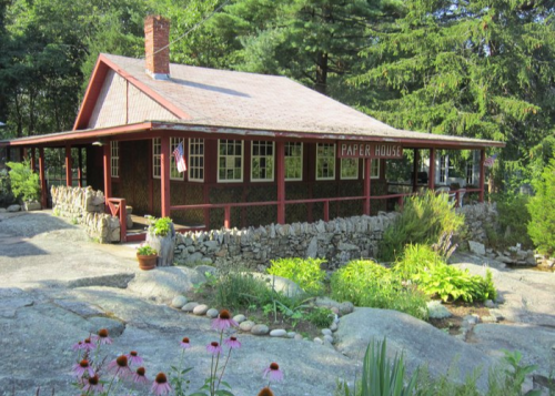A charming red house with a stone wall, surrounded by greenery and flowers, set in a peaceful outdoor environment.
