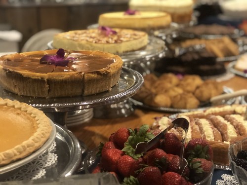 A variety of desserts on display, including cheesecakes, pies, and pastries, garnished with strawberries and flowers.