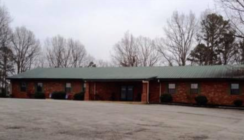 A single-story brick building with a green metal roof, surrounded by trees and a gravel parking lot.