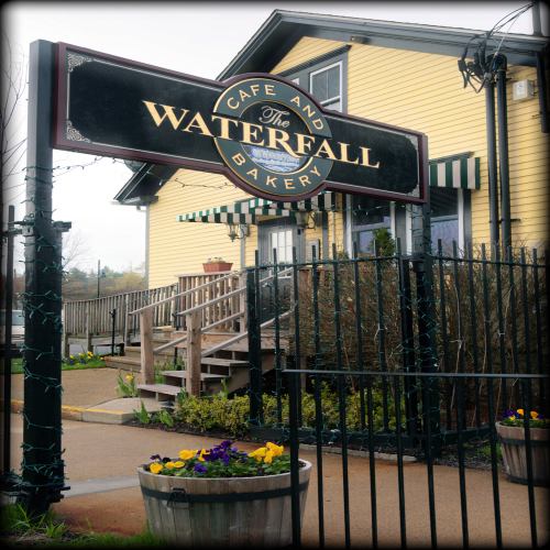 Sign for "The Waterfall Cafe and Bakery" in front of a yellow building with flowers and a fence.