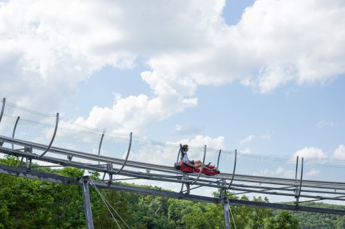 A person rides a summer coaster on a track above lush greenery under a partly cloudy sky.