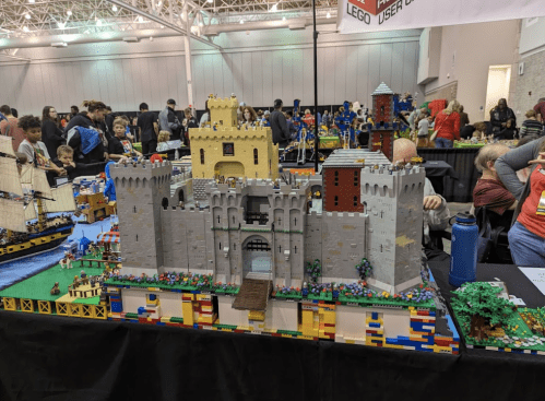 A detailed LEGO castle display with towers, a drawbridge, and colorful landscaping, surrounded by a crowd at an event.