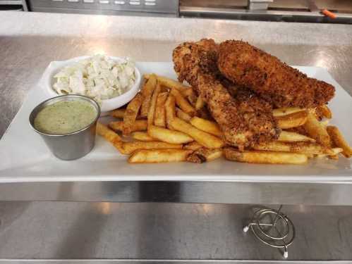 A plate of crispy fried chicken tenders, golden fries, coleslaw, and a small cup of green dipping sauce.