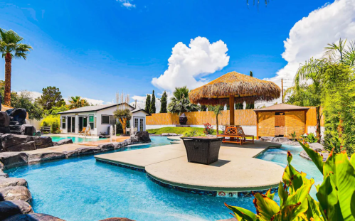 A sunny backyard with a pool, palm trees, a thatched gazebo, and a rock formation, surrounded by lush greenery.