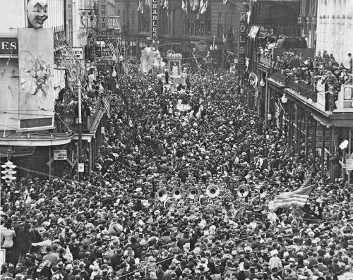 A large crowd gathers in a bustling street, filled with banners and decorations, during a festive parade.