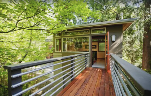 A modern cabin entrance surrounded by lush greenery, featuring a wooden deck and large windows.