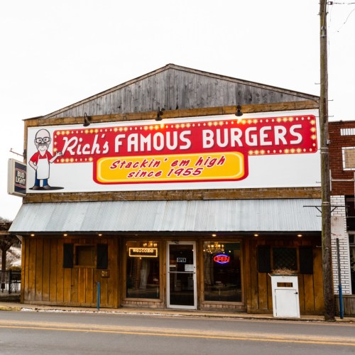A rustic burger restaurant with a large sign reading "Rich's Famous Burgers" and "Stackin' em high since 1955."