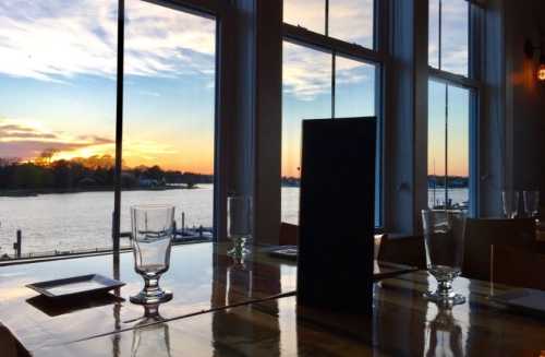 A restaurant table with glasses and a menu, overlooking a sunset by the water through large windows.