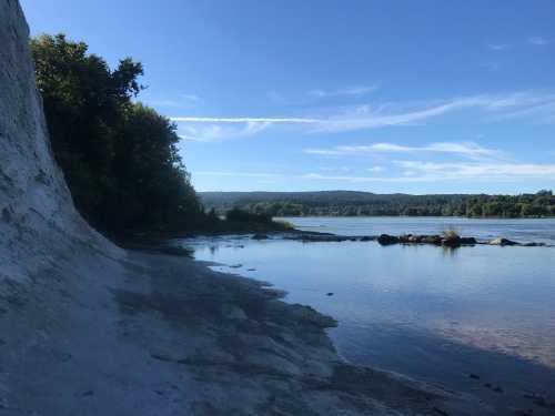 A serene riverside scene with a sandy shore, lush trees, and a clear blue sky reflecting on the water.