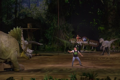 A performer interacts with life-sized dinosaur models on a stage set in a lush, prehistoric environment.