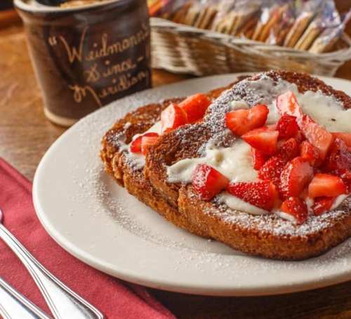 Plate of French toast topped with fresh strawberries and cream, dusted with powdered sugar, served with a coffee mug.