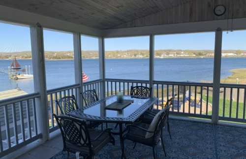 A screened porch with a dining table overlooks a calm lake and distant hills on a sunny day.