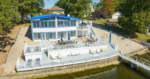 A modern waterfront home with a blue roof, large deck, and outdoor seating by the water, surrounded by trees.
