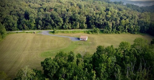 Aerial view of a grassy field with a small house surrounded by trees and a winding road in the background.