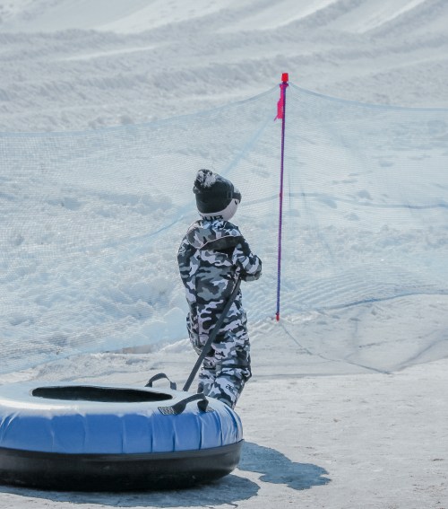 A child in a camouflage snowsuit stands near a snow-covered slope, holding a tube and looking towards a safety barrier.