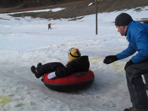 A child in a yellow hat sits on a red snow tube, while an adult helps them prepare to slide down a snowy hill.