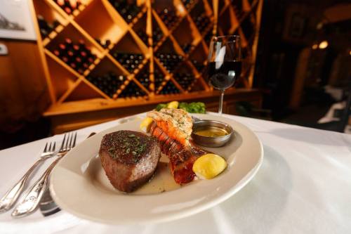A gourmet plate featuring steak, lobster, lemon, and a side of sauce, with a glass of red wine in a cozy setting.