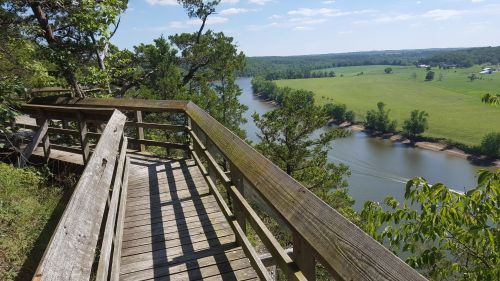 A wooden overlook with a railing, offering a view of a river and green fields under a clear blue sky.