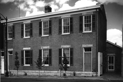 Black and white image of a two-story brick building with multiple windows and doors, surrounded by small trees.