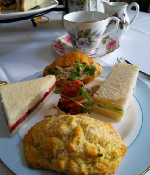 A plate with a scone, sandwich, croissant, and cucumber sandwich, with tea set in the background.