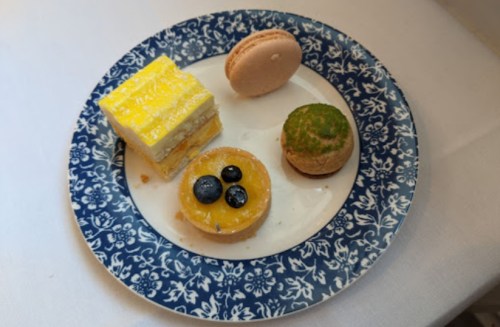 A blue floral plate with a slice of yellow cake, a macaron, a tart with blueberries, and a green-topped pastry.