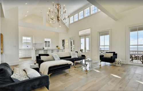 Bright, modern living room with large windows, elegant chandelier, and stylish black sofas on light wooden flooring.
