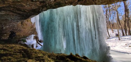 A frozen waterfall cascades down from a rocky overhang, surrounded by snow and trees under a clear blue sky.