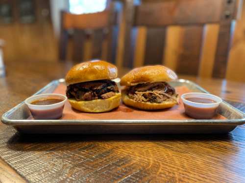 Two gourmet sandwiches on a tray, one with sliced meat and the other with pulled meat, accompanied by dipping sauces.