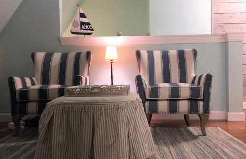 Two striped armchairs facing a small table with a lamp, set in a light green room with a nautical theme.