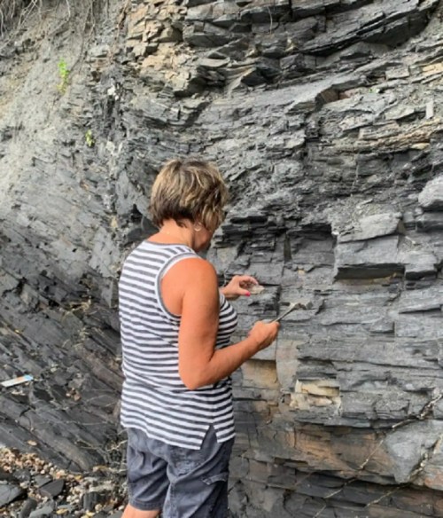 A woman examines rock layers on a cliffside, wearing a striped shirt and shorts, with tools in hand.
