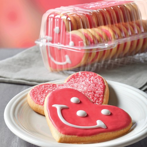 Two heart-shaped cookies with pink icing and smiley faces, placed on a white plate, with a container of cookies in the background.