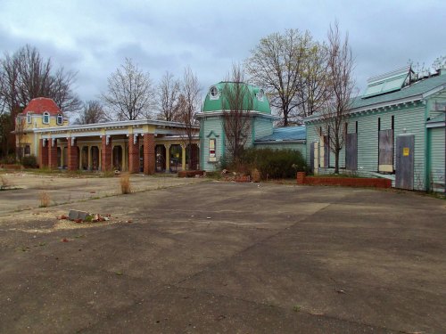 Abandoned building complex with a green dome, overgrown vegetation, and cracked pavement under a cloudy sky.
