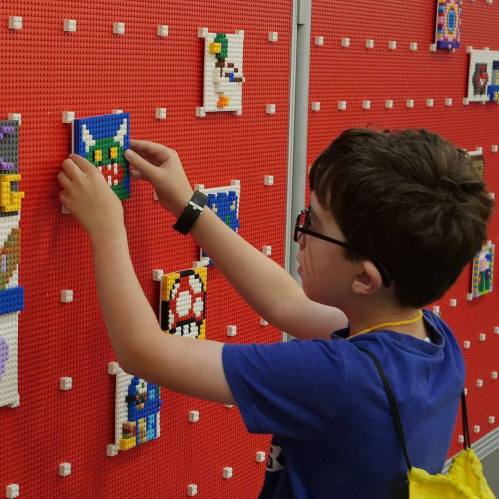 A child with glasses places a LEGO piece on a colorful display wall filled with various LEGO creations.