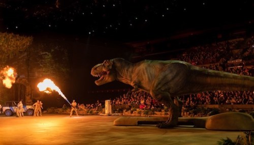 A large dinosaur performs on stage, with flames shooting from its mouth and a crowd watching in the background.