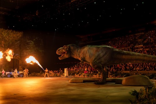 A lifelike dinosaur performs on stage, breathing fire, with a large audience watching in an arena setting.