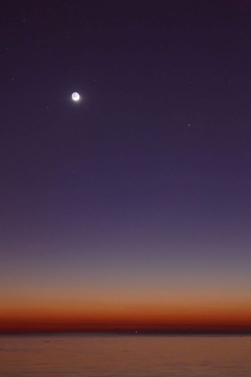 A serene twilight sky with a crescent moon and stars above a calm ocean, transitioning from deep blue to warm orange hues.