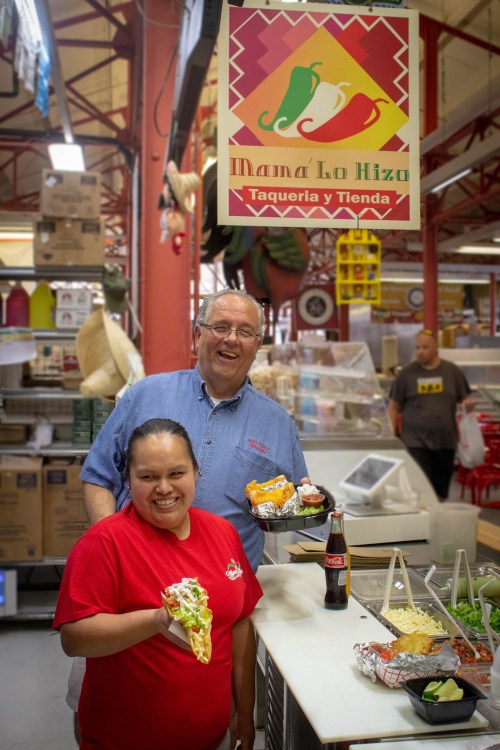 A smiling woman holds a plate of food in a market, while a man stands behind her, both in front of a taqueria sign.
