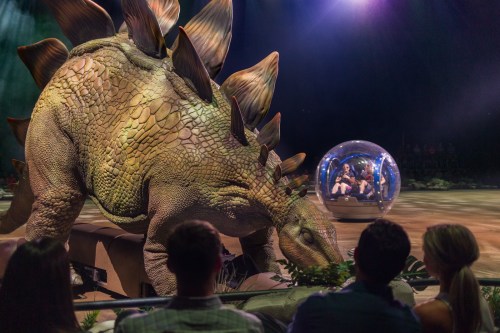 A large dinosaur model interacts with a transparent bubble containing people, with an audience watching in the background.