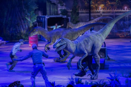 A dramatic scene with lifelike dinosaur models and performers in a colorful, staged environment.
