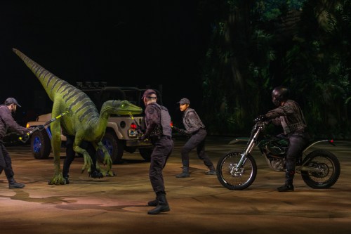 A green dinosaur is being controlled by performers in black outfits, with a motorcycle and vehicle in the background.