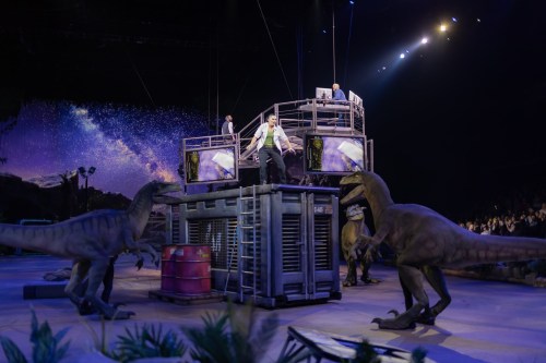 A dramatic scene with actors on a platform surrounded by life-sized dinosaur models in a theatrical performance.